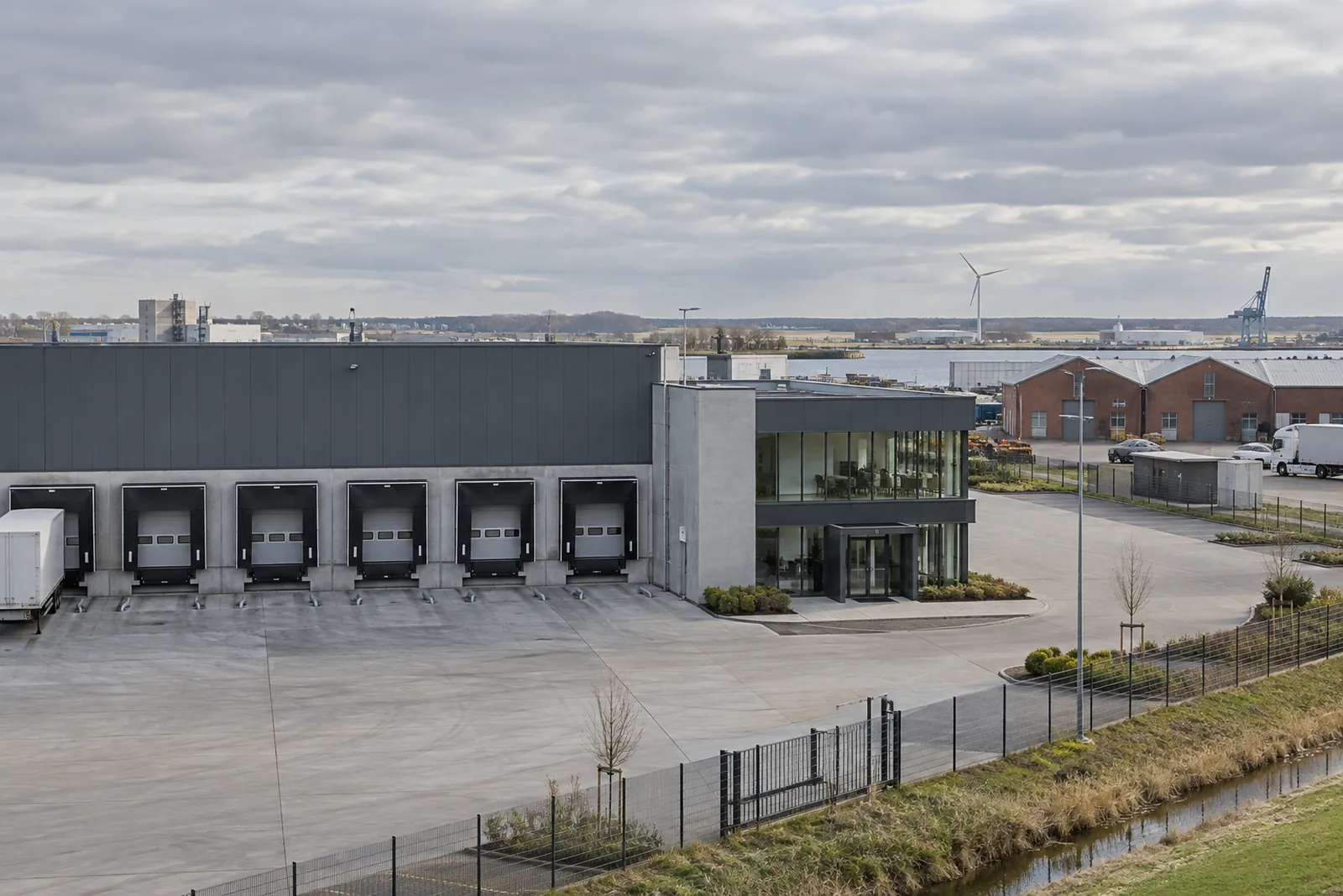 Lagerhalle mit Ladetoren und verglastem Büroanbau in einem Gewerbegebiet im norddeutschen Flachland, Windrad am Horizont
