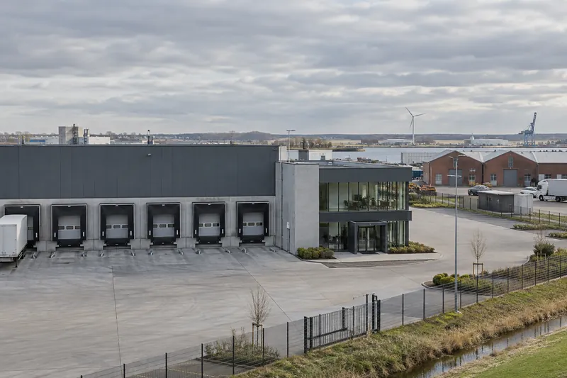 Lagerhalle mit Ladetoren und verglastem Büroanbau in einem Gewerbegebiet im norddeutschen Flachland, Windrad am Horizont