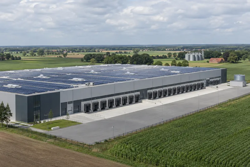 Logistikhalle mit Solardach auf flachem Land im Oldenburger Münsterland, im Hintergrund Silos und ein Backsteinhof