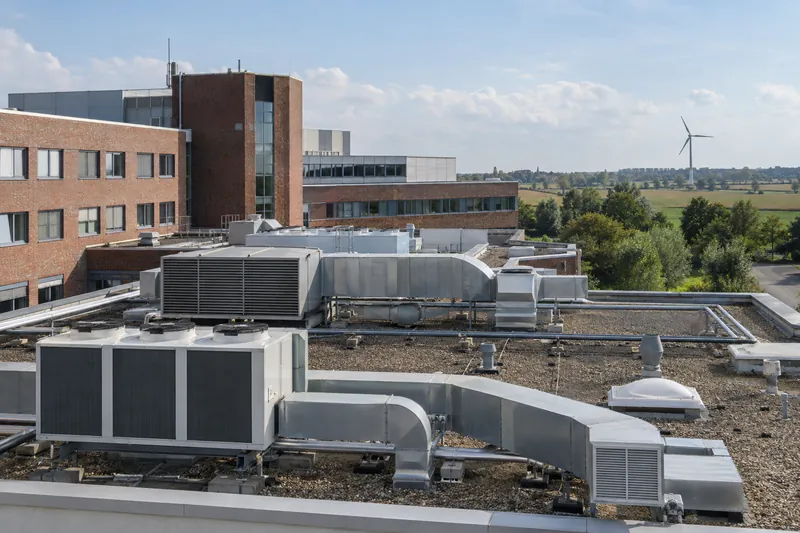 Klimageräte und Lüftungskanäle auf dem Flachdach eines Gewerbebaus, dahinter Klinkerfassade und Windrad am Horizont