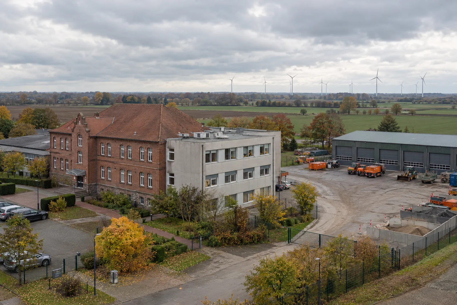 Backstein-Kreishaus mit Anbau aus den 1970er Jahren, daneben die Stahlhalle eines Bauhofs, dahinter flaches Land mit Windrädern