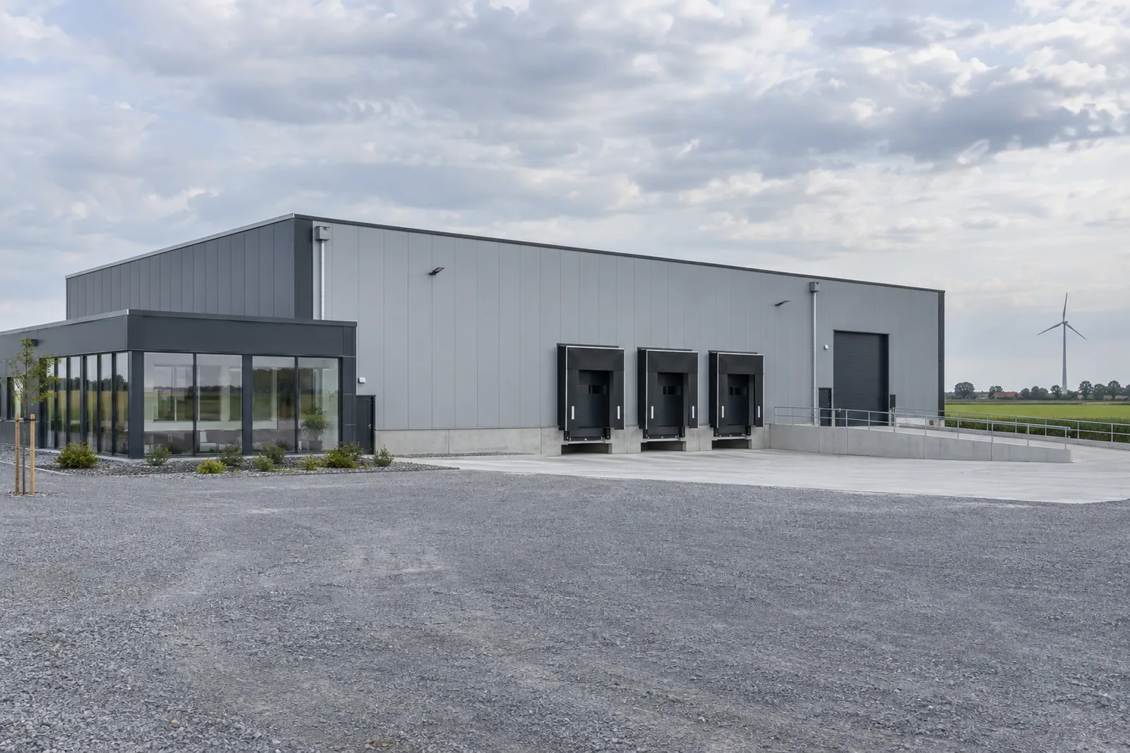 Neu errichtete Logistikhalle mit kleinem Bürotrakt auf flachem Land in Niedersachsen, Windrad am Horizont