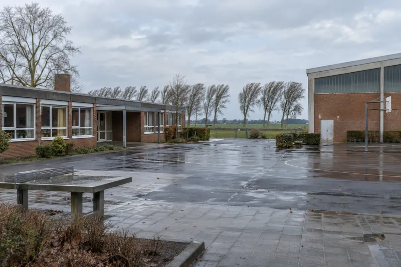 Backsteinschule der 1970er Jahre mit Klassentrakt und Turnhalle am Ortsrand, dahinter flaches Land mit Baumreihe