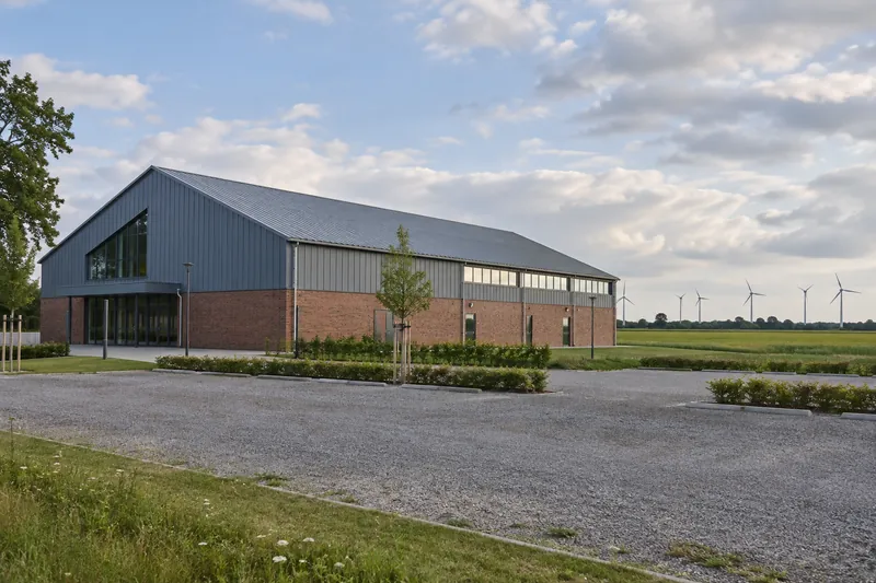 Mehrzweck-Sporthalle mit Backsteinsockel und Metalldach am Ortsrand, dahinter flaches Land und Windräder am Horizont