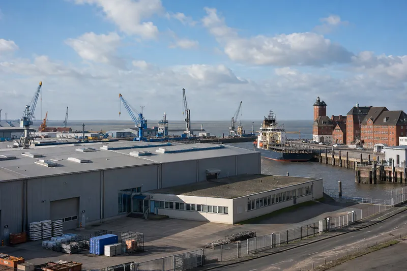 Lagerhalle mit Bürobau im Hafen von Cuxhaven, dahinter Kräne, ein Schiff und die offene Elbmündung