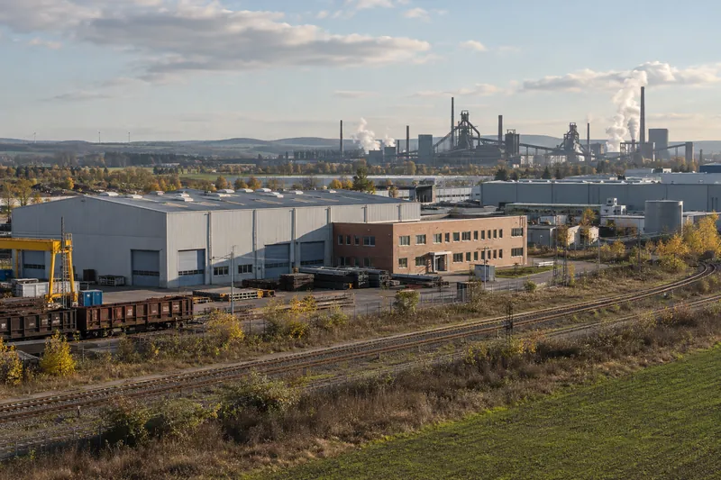 Produktionshalle mit Bürotrakt, im Hintergrund die Silhouette eines Stahlwerks bei Salzgitter