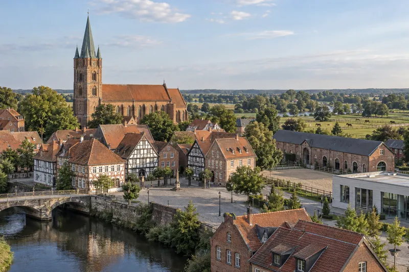 Fachwerk- und Backsteinhäuser vor einem Dom mit Backsteinturm, am Stadtrand eine Reithalle und Weiden